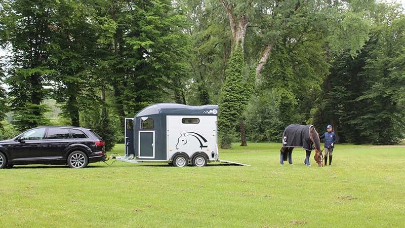 Uma carrinha atrelada a um carro num prado verde. Um cavalo e um cavaleiro pastam nas proximidades.