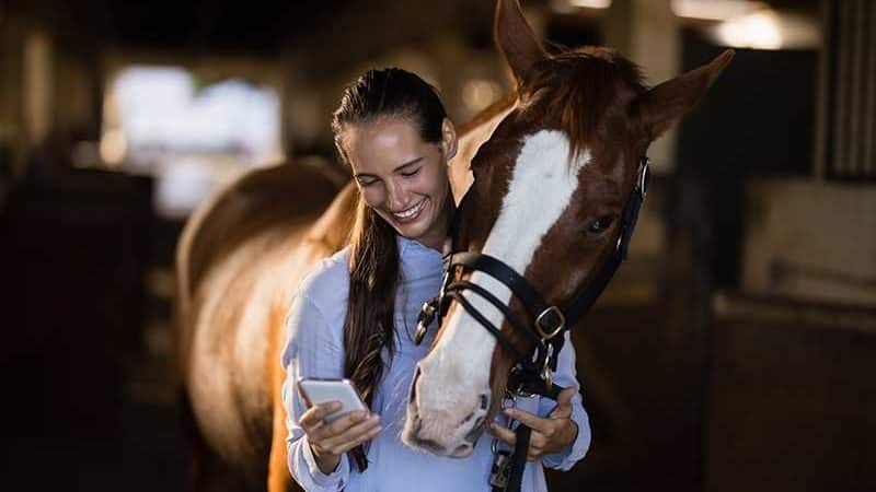 Mulher jovem a olhar para o seu smartphone e a sorrir. O cavalo está a olhar por cima do ombro.