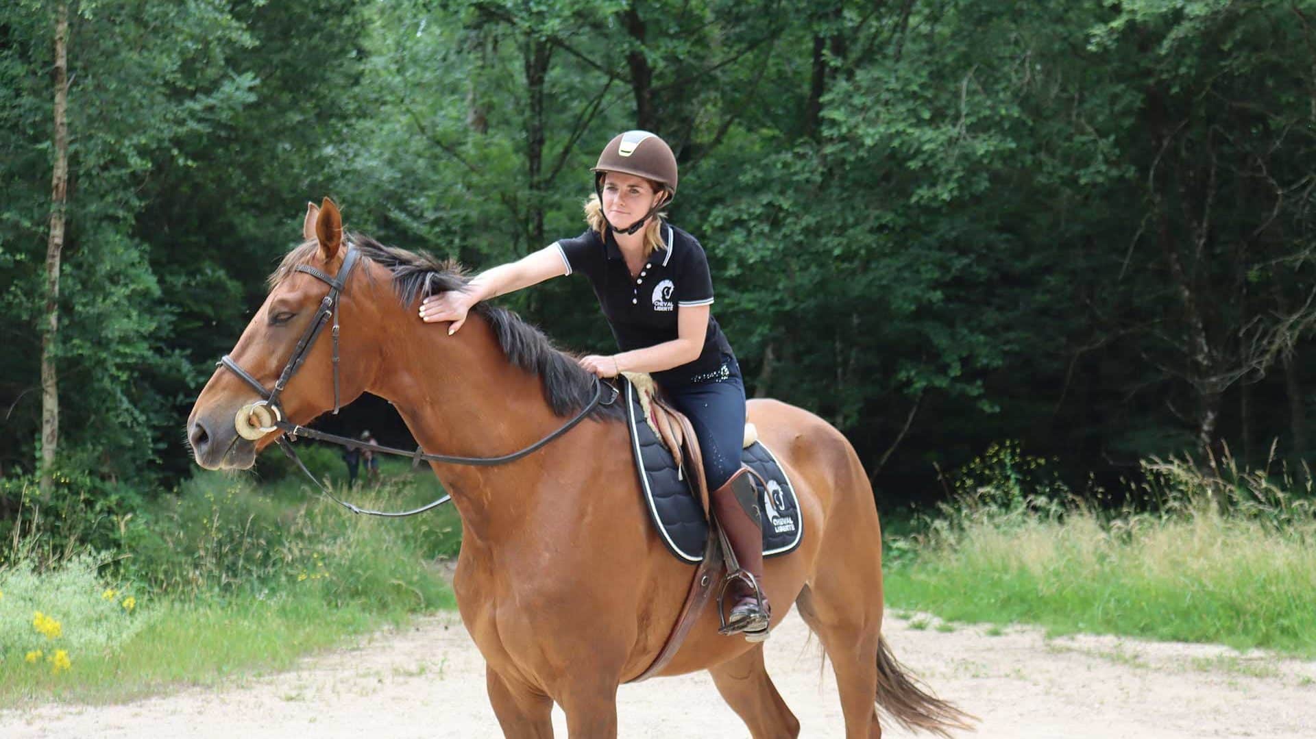 Uma cavaleira parada acaricia o seu cavalo na floresta, sorrindo.